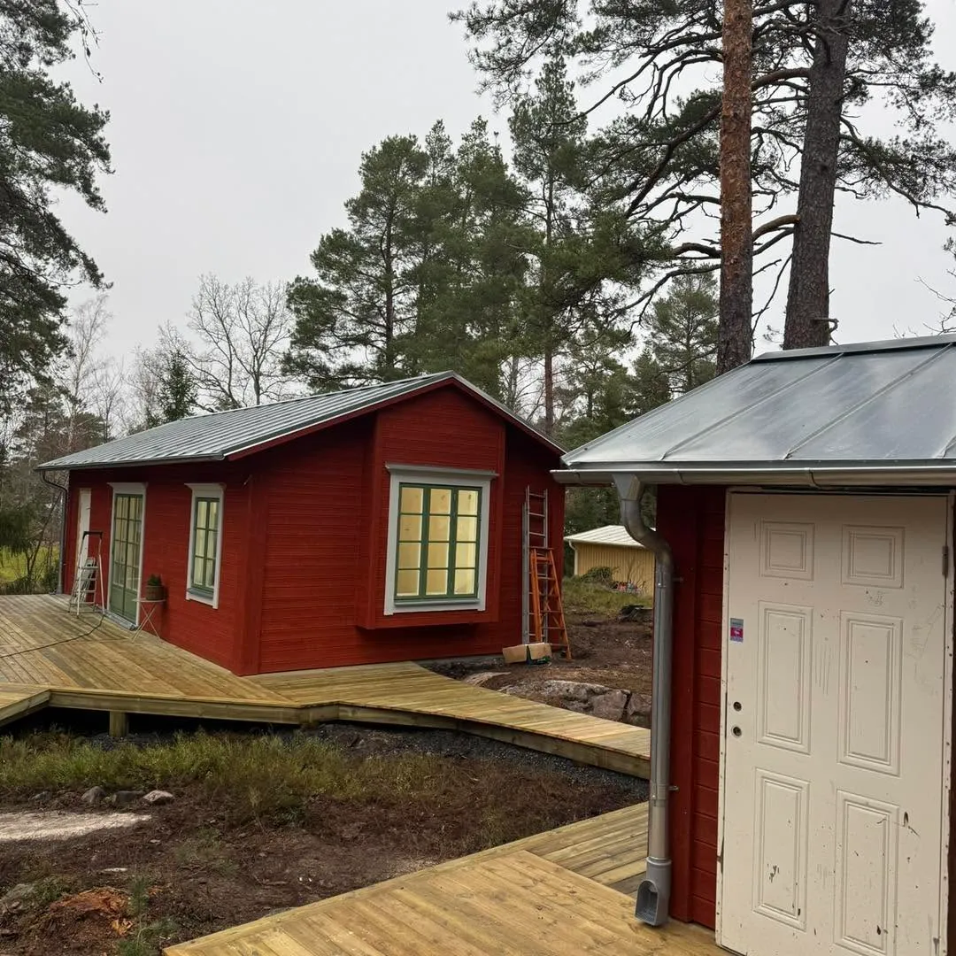 Röd stuga med plåttak och vitt uthus i skogen
